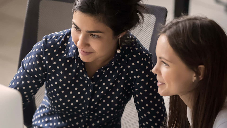 Multi ethnic female colleagues sit at workplace working together looks at pc screen use corporate program, apprentice listen mentor, teamwork concept.