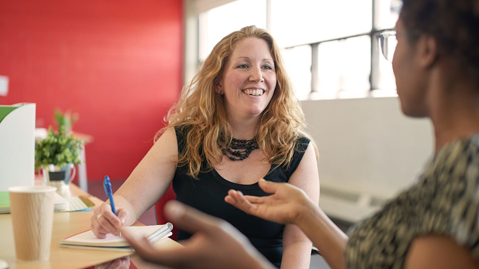 Woman chatting to colleague and writing in a notebook